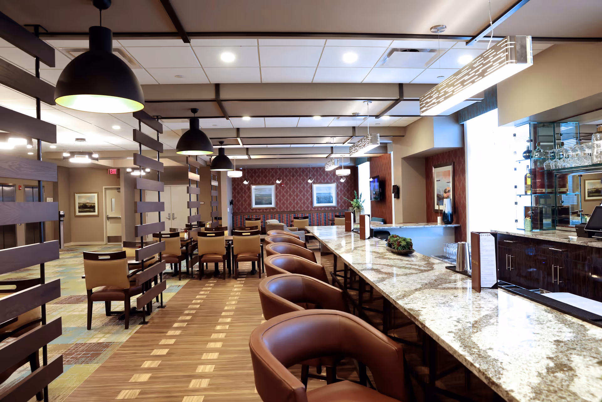Interior view of a dining area in a retirement community featuring a long granite countertop with brown leather bar stools, multiple tables with chairs, modern pendant lighting, and decorative wall art in the background.