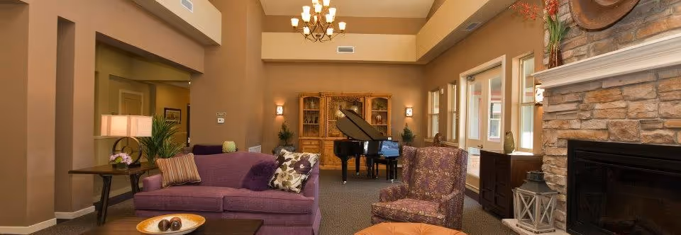 A cozy living room area in Deer Trail Assisted Living featuring a purple sofa with decorative pillows, a patterned armchair, a wooden coffee table with a decorative bowl, a stone fireplace with a lantern and floral arrangement, a grand piano in the background, warm beige walls, and large windows letting in natural light.