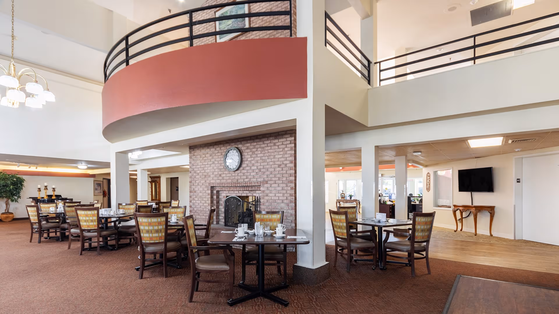 Spacious dining area with multiple tables and chairs arranged around a brick fireplace with a clock above it. The room features high ceilings with a balcony railing above, a chandelier, and a mix of carpet and wood flooring. The space is well-lit with natural light coming from windows in the background.