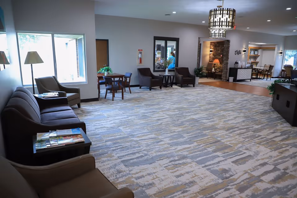 A spacious and well-lit common area in an assisted living facility featuring multiple seating arrangements including sofas, armchairs, and small tables. The room has large windows allowing natural light, a patterned carpet, modern light fixtures, and decorative plants. There is a cozy nook with a lamp and bookshelves in the background.