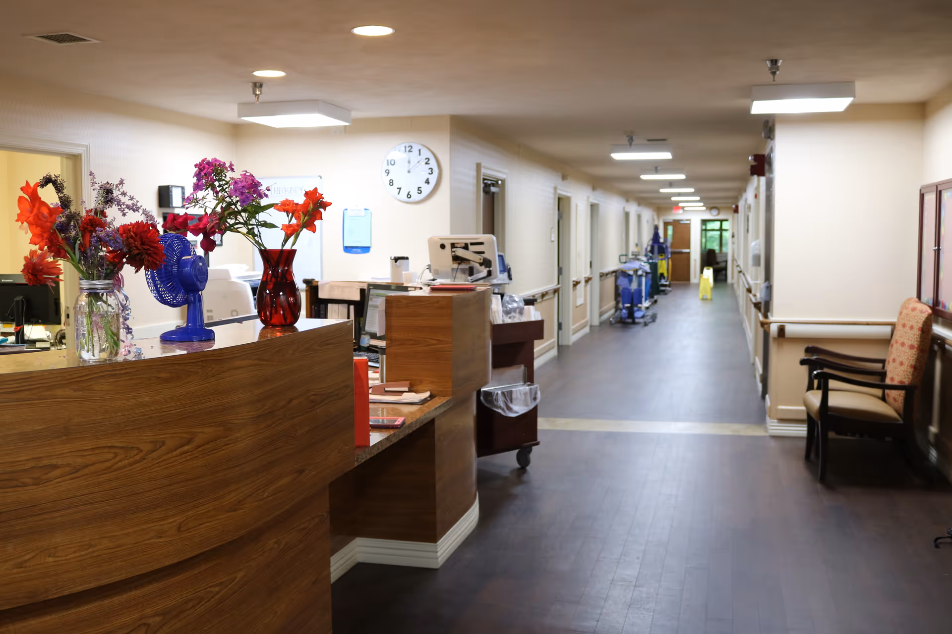 Interior view of a senior living facility hallway with a wooden reception desk on the left adorned with vases of colorful flowers and a blue fan. The hallway has multiple doors on both sides, cleaning carts further down the corridor, a clock on the wall, and a chair with patterned upholstery on the right side.