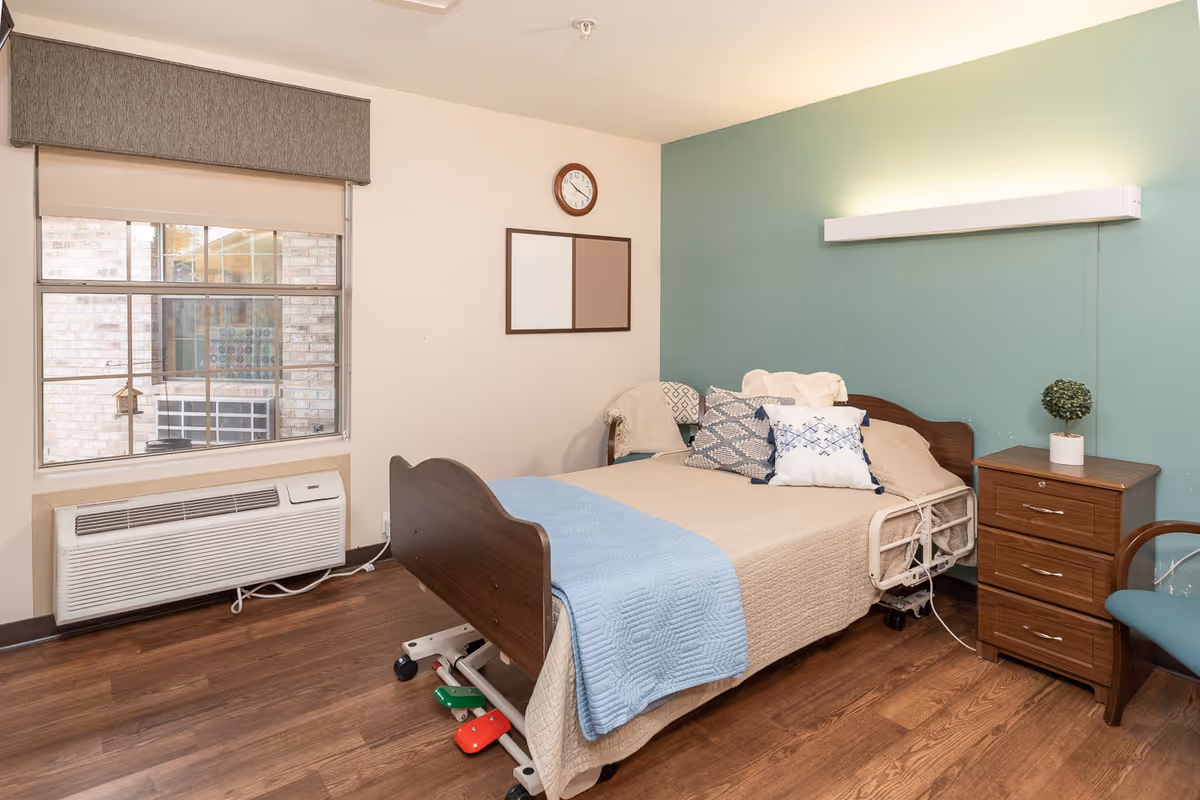 A nursing facility bedroom with a hospital-style bed covered with beige and blue bedding, several decorative pillows, a wooden nightstand with a small potted plant, a chair with a cushion, a window with a gray valance, a wall clock, and a bulletin board on the wall.