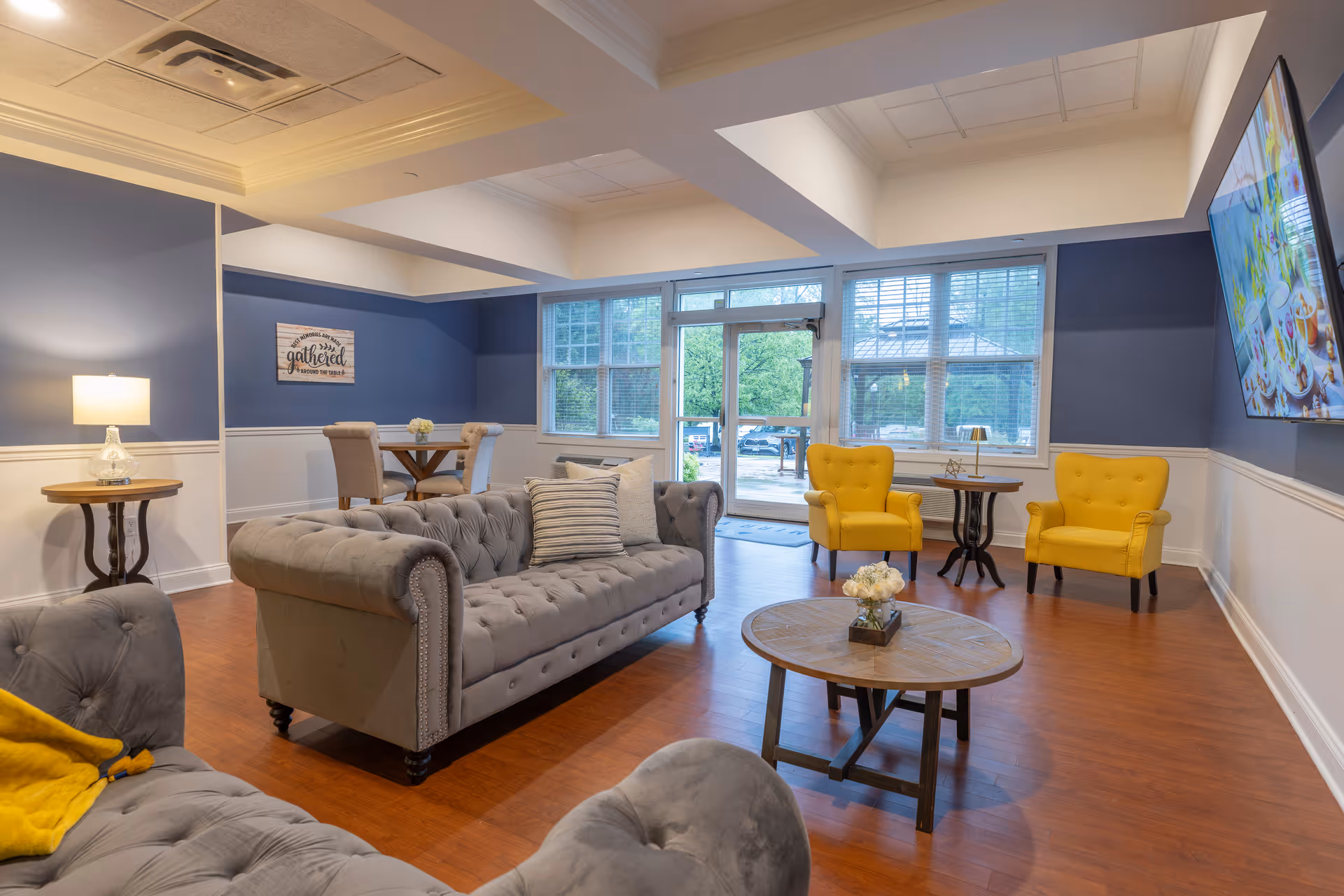 A bright and welcoming living room area with blue and white walls, wooden flooring, and large windows letting in natural light. The room features two gray tufted sofas, two yellow armchairs, a round wooden coffee table with a flower arrangement, a side table with a lamp, and a wall-mounted TV displaying a colorful image. There is also a small dining table with chairs in the corner and a decorative sign on the wall.