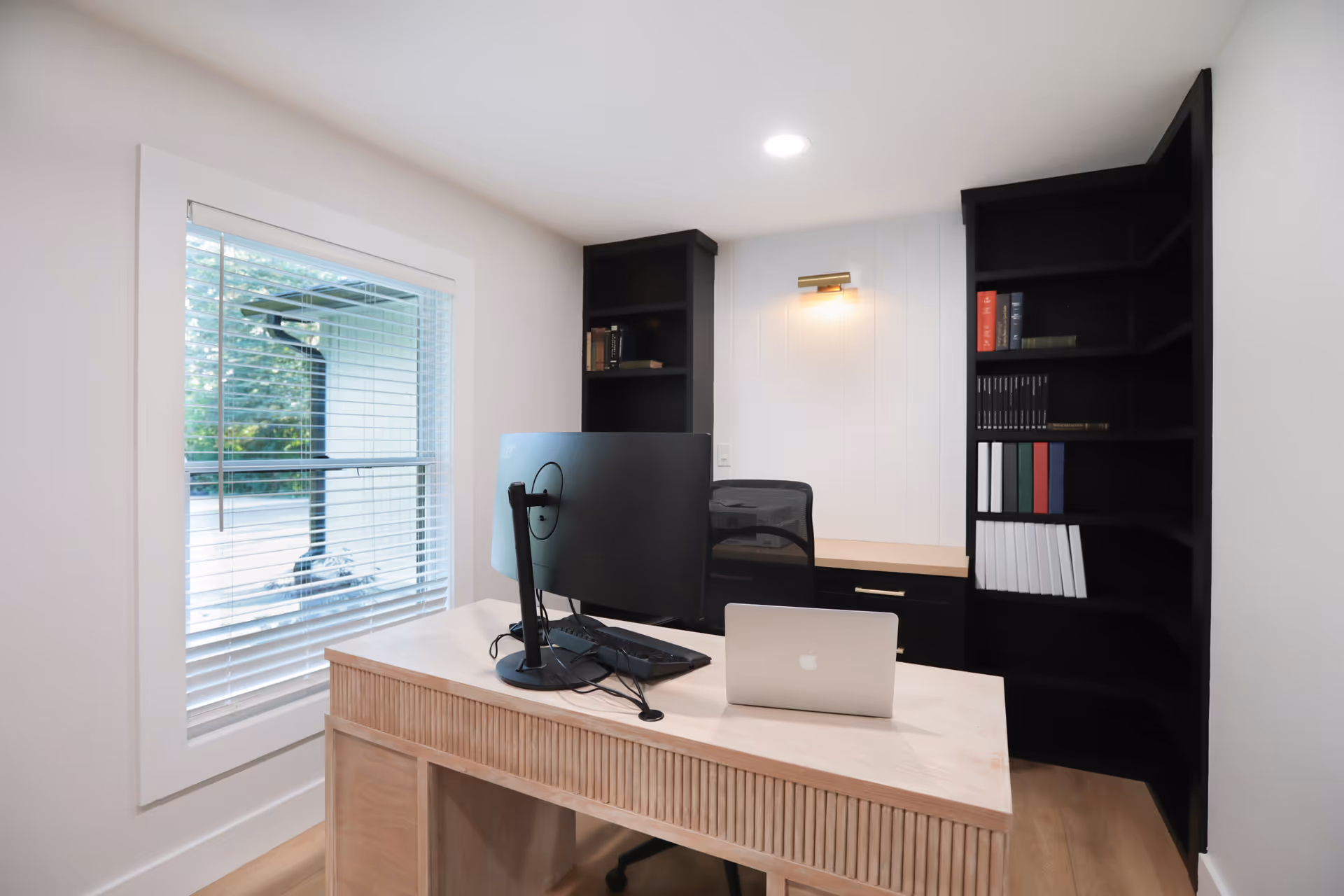 Small home office with a wooden desk, computer monitors and laptop, black bookshelves, and a window with blinds.