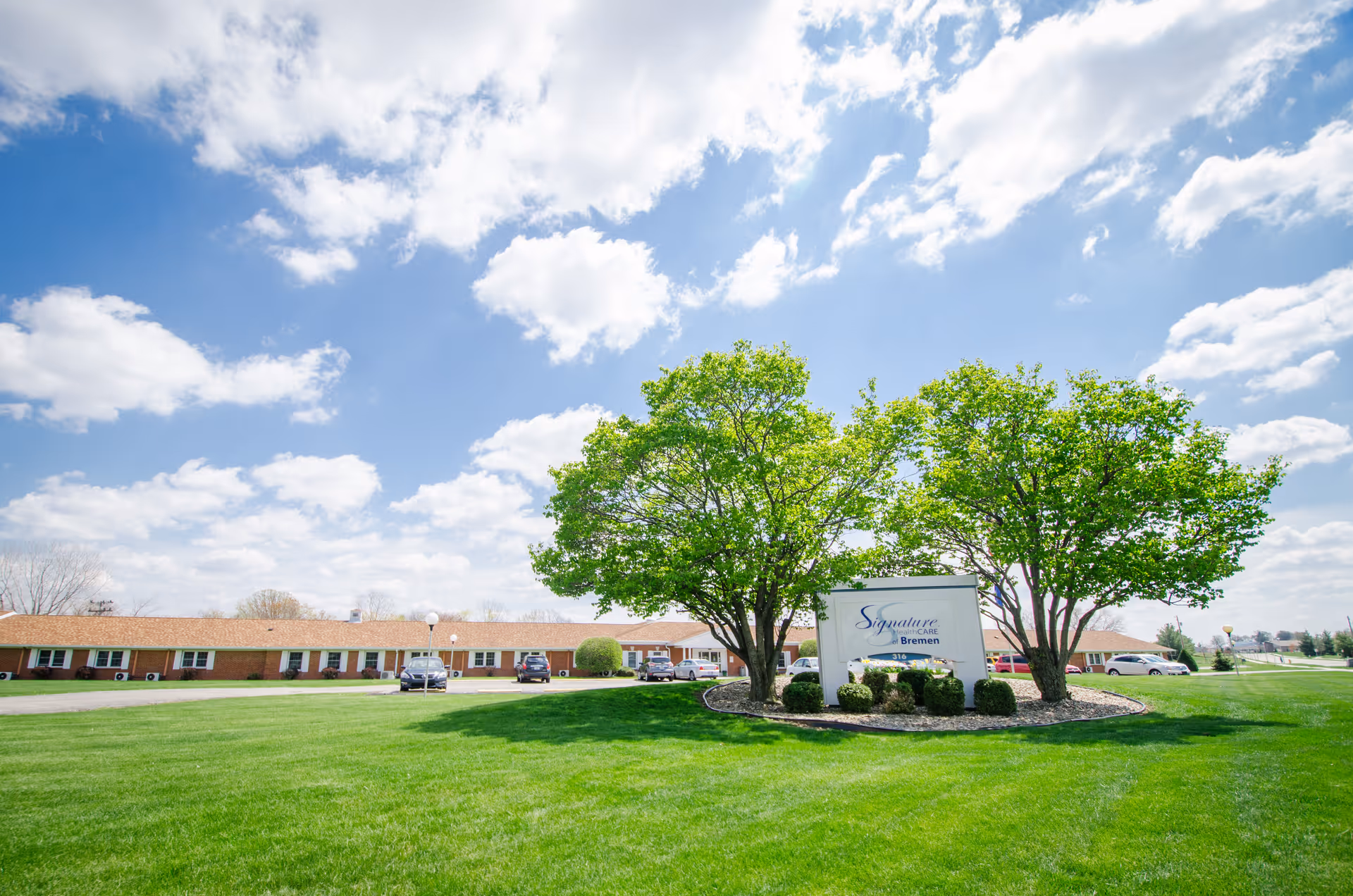 Front lawn and sign for Signature HealthCARE of Bremen with a low brick building, trees, and a blue sky.