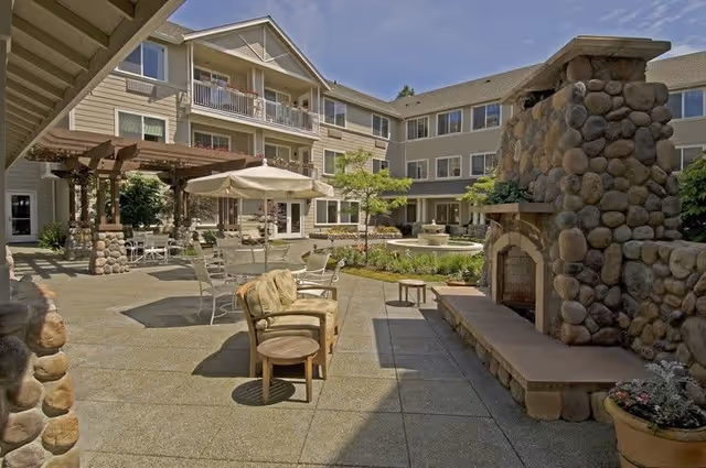 Outdoor courtyard area of a senior living facility with stone fireplace, patio seating including chairs and tables with umbrellas, a pergola, and a three-story building surrounding the courtyard.