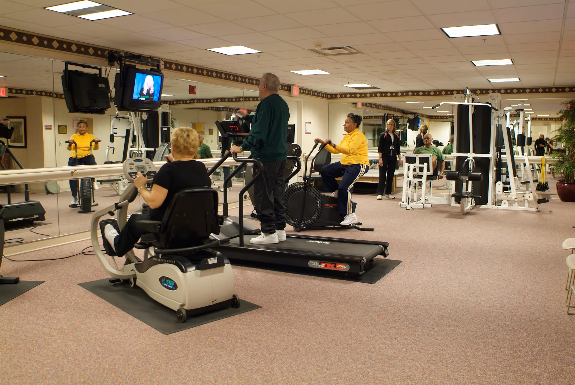 A fitness room in Eastcastle Place, a CCRC, with several seniors exercising on various gym equipment including a recumbent bike, treadmill, and stationary bike. A TV is mounted on the wall, and a staff member is standing near the exercise machines. The room has carpeted floors, mirrored walls, and exercise machines in the background.