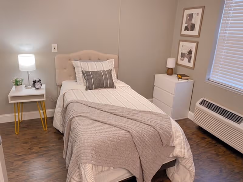 A neatly made single bed with a beige tufted headboard, white and gray striped bedding, and a gray throw blanket in a cozy room. To the left of the bed is a small white nightstand with yellow legs, holding a lamp, a small plant, and an alarm clock. To the right is a white dresser with a small lamp and books on top. The room has wooden flooring, light gray walls, two framed abstract artworks on the wall, a window with blinds, and a wall-mounted air conditioning unit below the window.