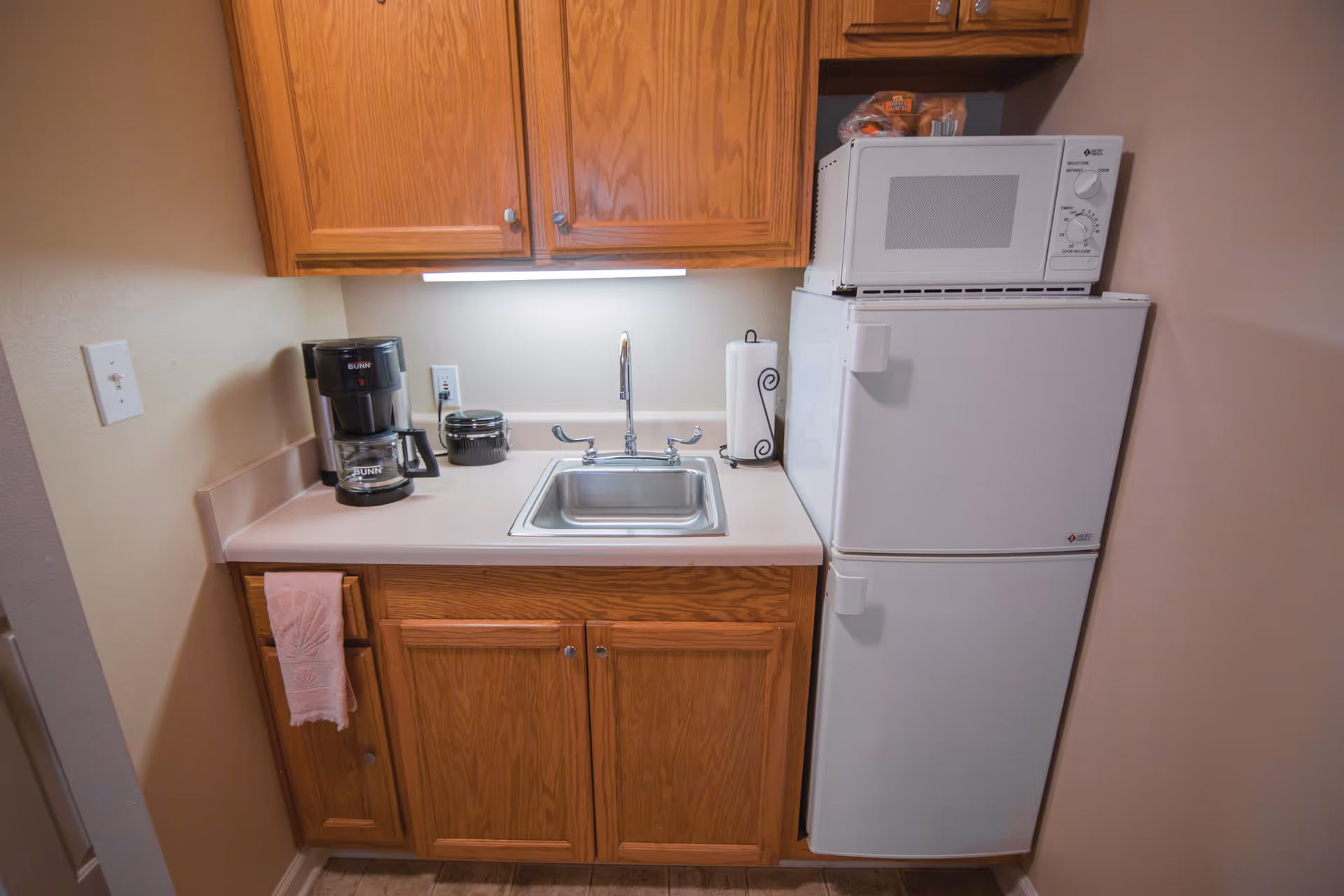 Small kitchenette with wooden cabinets, a sink, coffee maker, microwave, and a compact refrigerator.