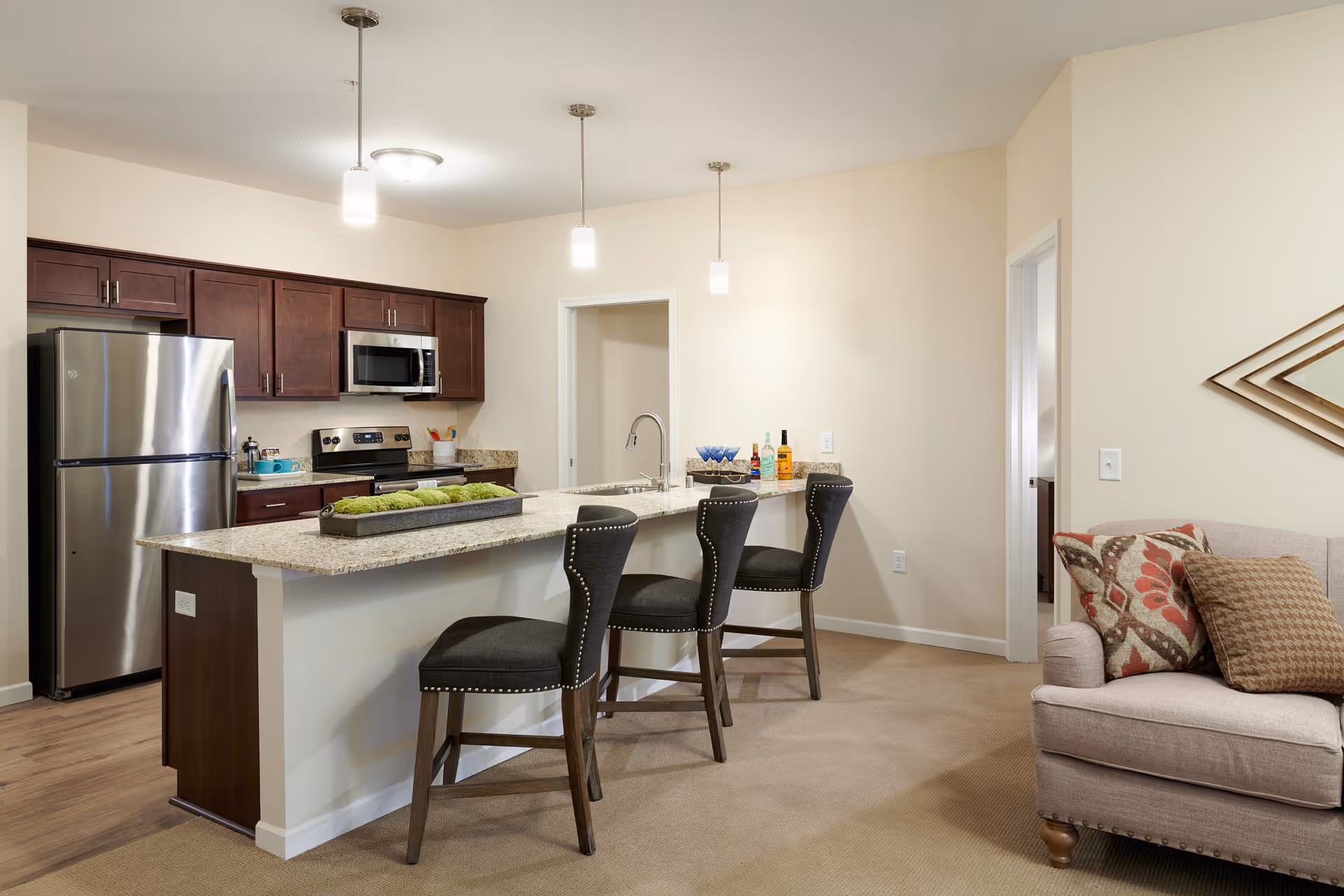 A modern kitchen area with dark wood cabinets, stainless steel refrigerator, stove, and microwave. A granite countertop island with three black cushioned bar stools is in the foreground. The kitchen opens into a living space with a beige armchair adorned with patterned pillows. The walls are light-colored, and three pendant lights hang above the island.