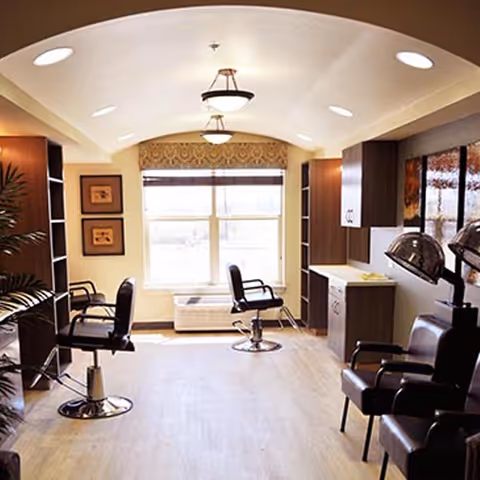 Interior of a salon area in a senior living facility with salon chairs, hair drying stations, wooden cabinets, and a large window letting in natural light.