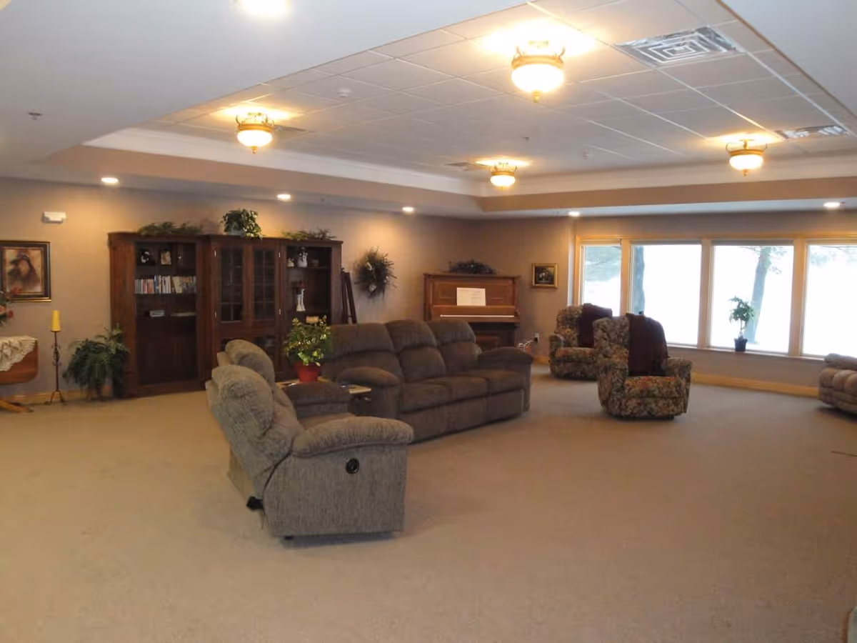 A spacious living room with beige carpet and neutral walls, featuring a gray reclining sofa set, two floral armchairs, a wooden cabinet with glass doors, a piano, and large windows letting in natural light. The ceiling has recessed lighting and decorative light fixtures.