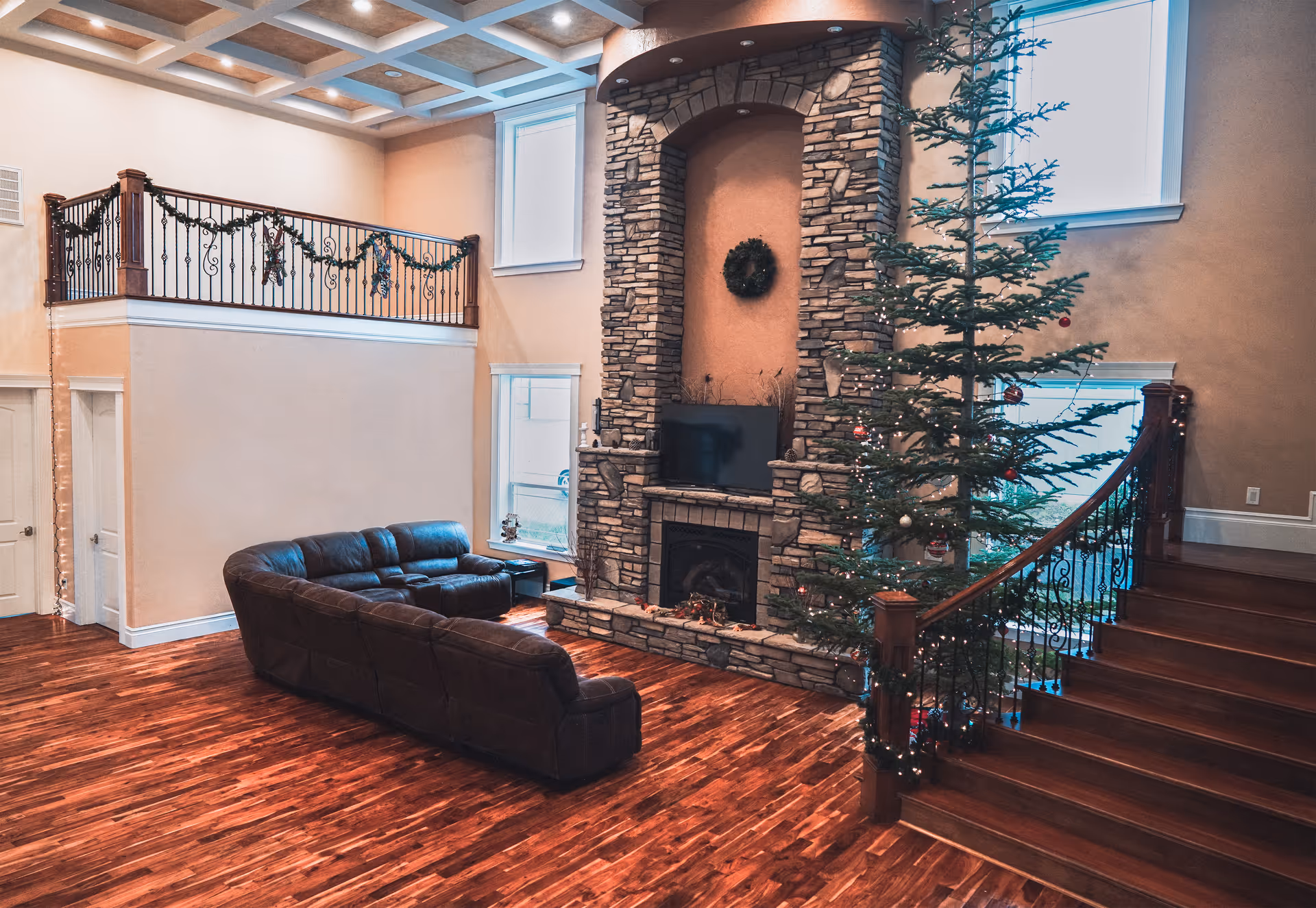 Spacious living room with wooden flooring, a large stone fireplace with a TV mounted above it, a dark brown leather sectional sofa, a decorated Christmas tree, and a staircase with ornate railing. The room has high ceilings with recessed lighting and two large windows.