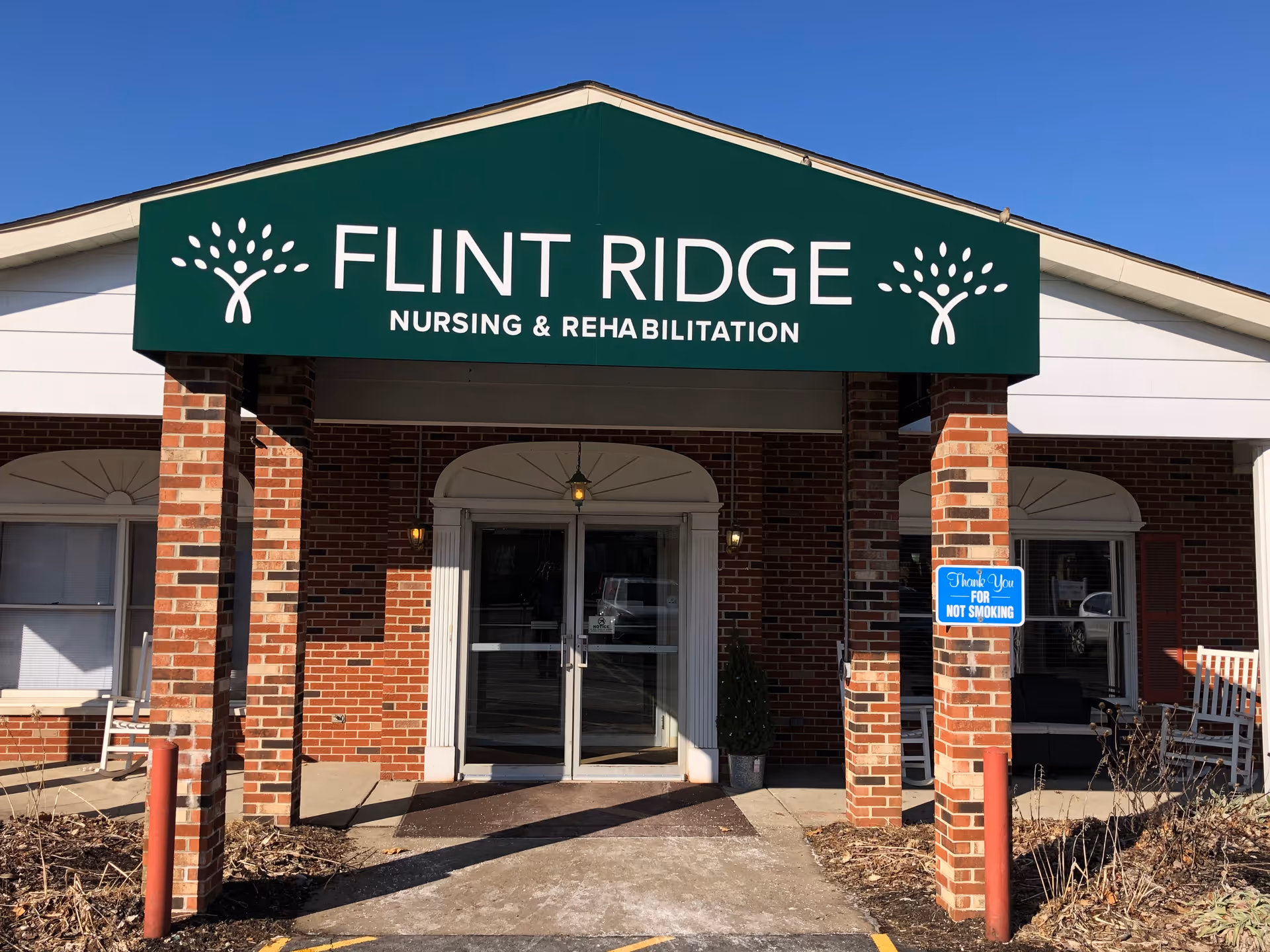 Entrance of Flint Ridge Nursing and Rehabilitation facility with a green awning displaying the facility name and logo. The building has red brick pillars and walls, glass double doors, and a 'Thank You For Not Smoking' sign on one pillar. There are benches and some plants near the entrance.