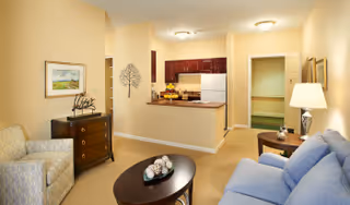 A cozy living room area in a senior living facility with a blue sofa, a wooden coffee table with decorative spheres, a side table with a lamp, and a chest of drawers with a framed painting and decorative items. The room opens into a kitchen area with dark wood cabinets, a white refrigerator, and a countertop with fruit bowls. The walls are painted a warm beige color.