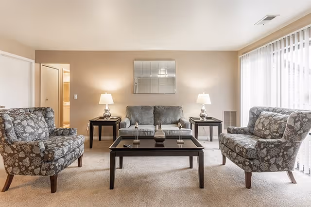 A living room with a gray sofa and two floral patterned armchairs arranged around a dark wooden coffee table. Two matching side tables with lamps flank the sofa. A large window with vertical blinds allows natural light to fill the room. A square mirror hangs on the beige wall behind the sofa.