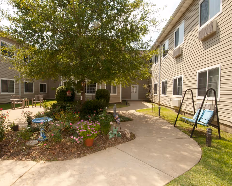 Sunlit courtyard of a senior living facility with a central tree and flower garden, paved walkway, benches and a blue porch swing between two building wings.