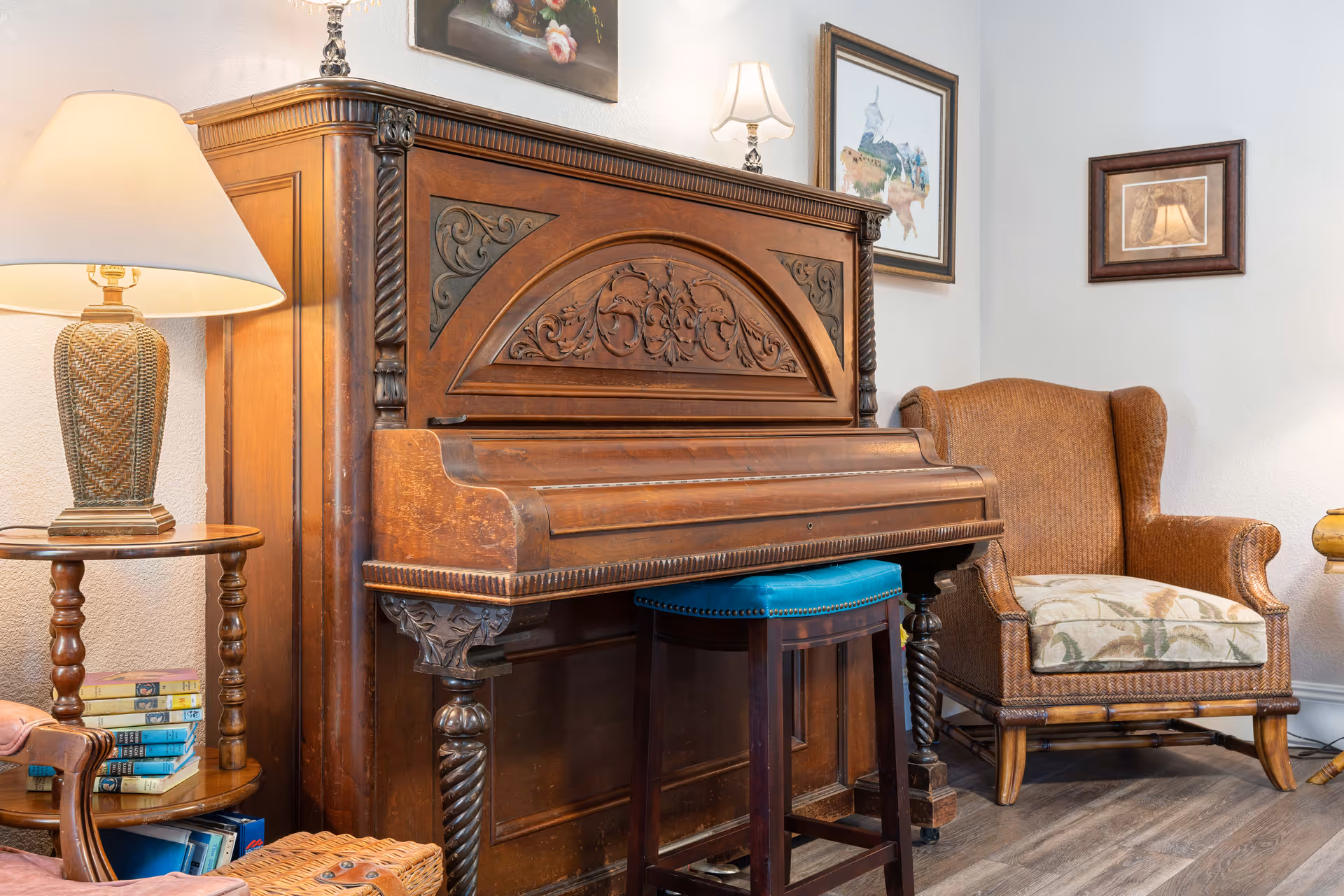 A cozy living room corner featuring an ornately carved wooden upright piano with a blue cushioned stool in front. To the right is a brown upholstered armchair with a floral cushion. On the left, there is a wooden side table with a lamp and a stack of books. The walls have framed artwork and the floor is wood.