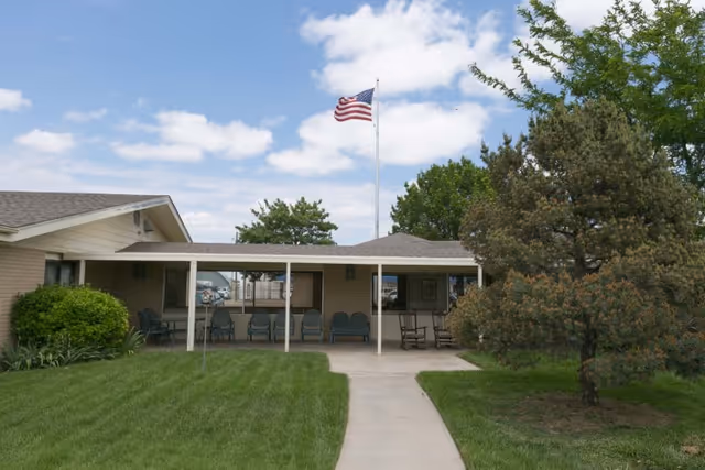 Exterior view of a single-story building with a covered porch area featuring several chairs. A concrete walkway leads to the porch, surrounded by green grass and trees. An American flag flies on a flagpole in front of the building under a partly cloudy sky.