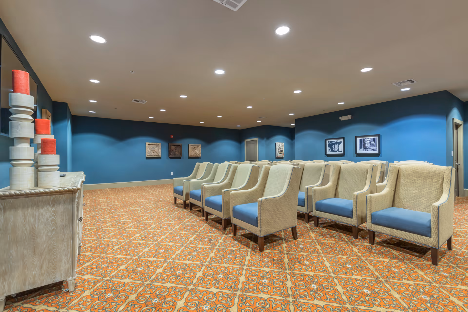 A room with rows of beige armchairs with blue cushions arranged facing forward on a patterned orange and brown carpet. The walls are painted blue and decorated with framed artwork. On the left side, there is a wooden cabinet with stacked decorative candles. The ceiling has multiple recessed lights.