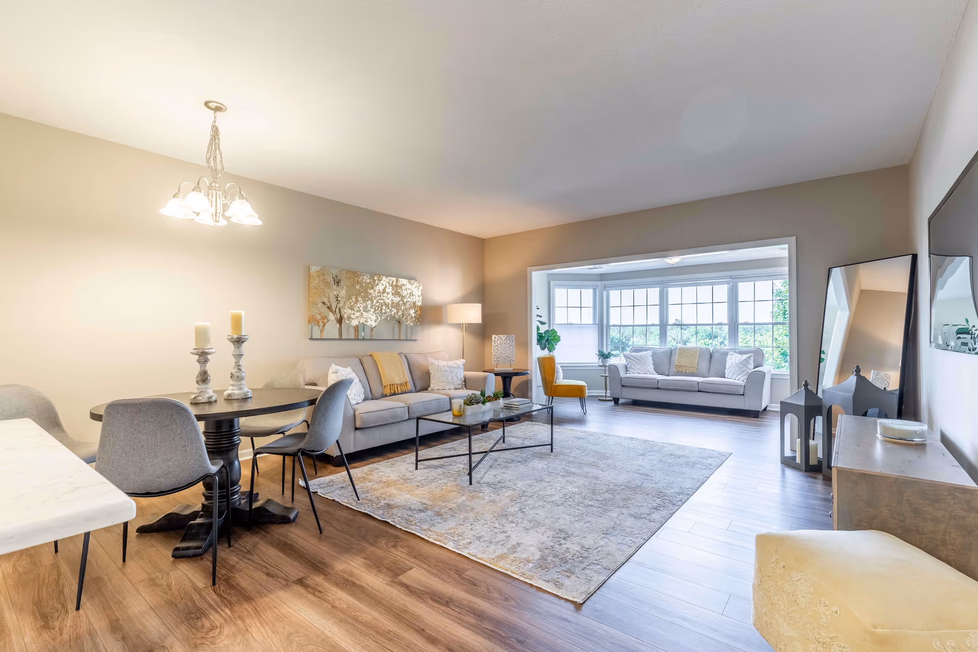 A bright and spacious living room and dining area with wooden flooring. The dining area features a round table with four gray chairs and two candle holders. The living room has a large area rug, a coffee table, a gray sofa with cushions, a floor lamp, and a yellow accent chair. In the background, there is a sunroom with large windows, a gray sofa, and plants. A large mirror and a TV are visible on the right side of the room.