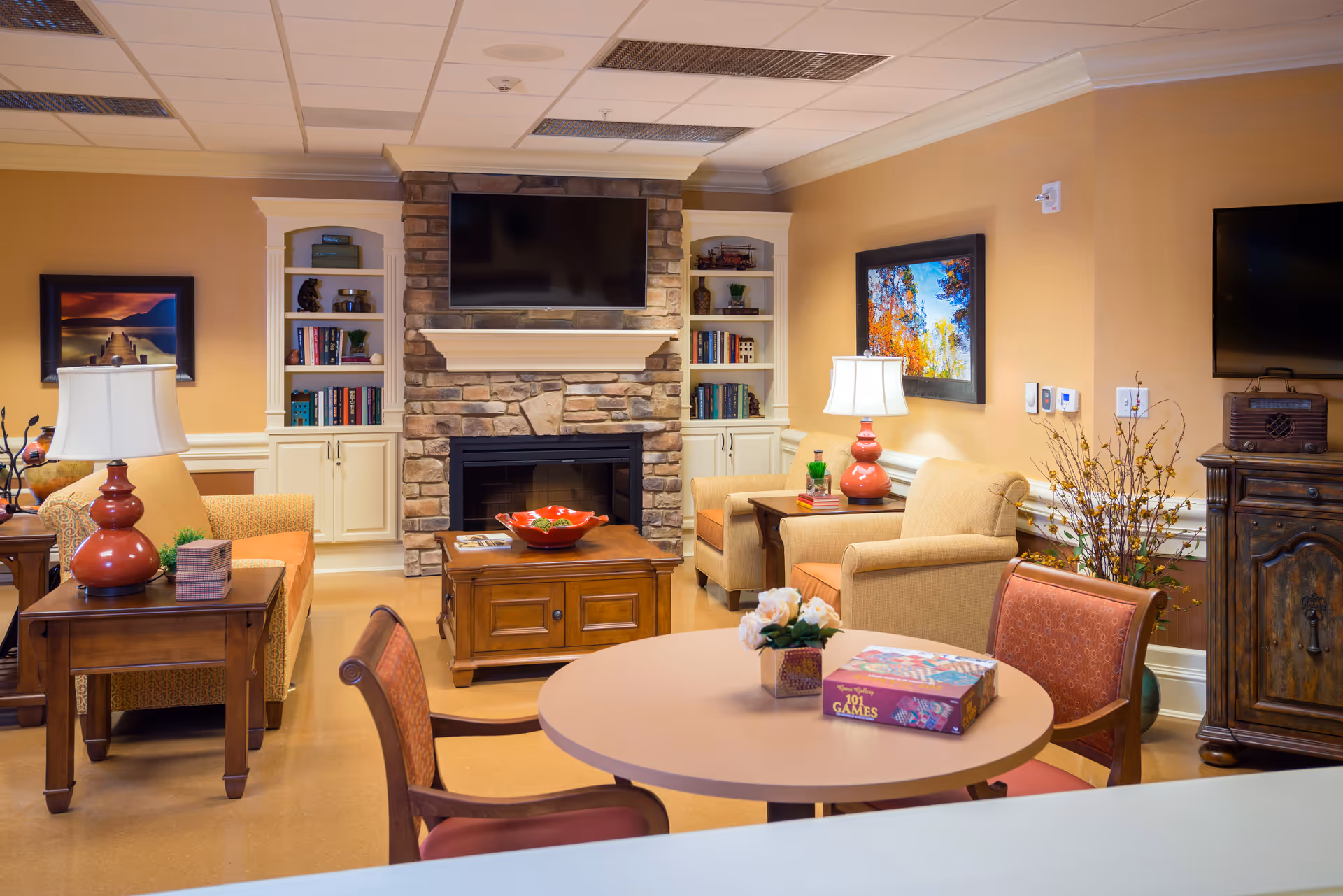 A cozy living room area in a senior living facility featuring comfortable armchairs and sofas arranged around a wooden coffee table with a decorative bowl. The room has a stone fireplace with a mounted flat-screen TV above it, built-in white bookshelves filled with books and decor, and warm beige walls adorned with colorful framed artwork. A round table with chairs and a box of games is in the foreground, along with table lamps and floral arrangements adding to the inviting atmosphere.
