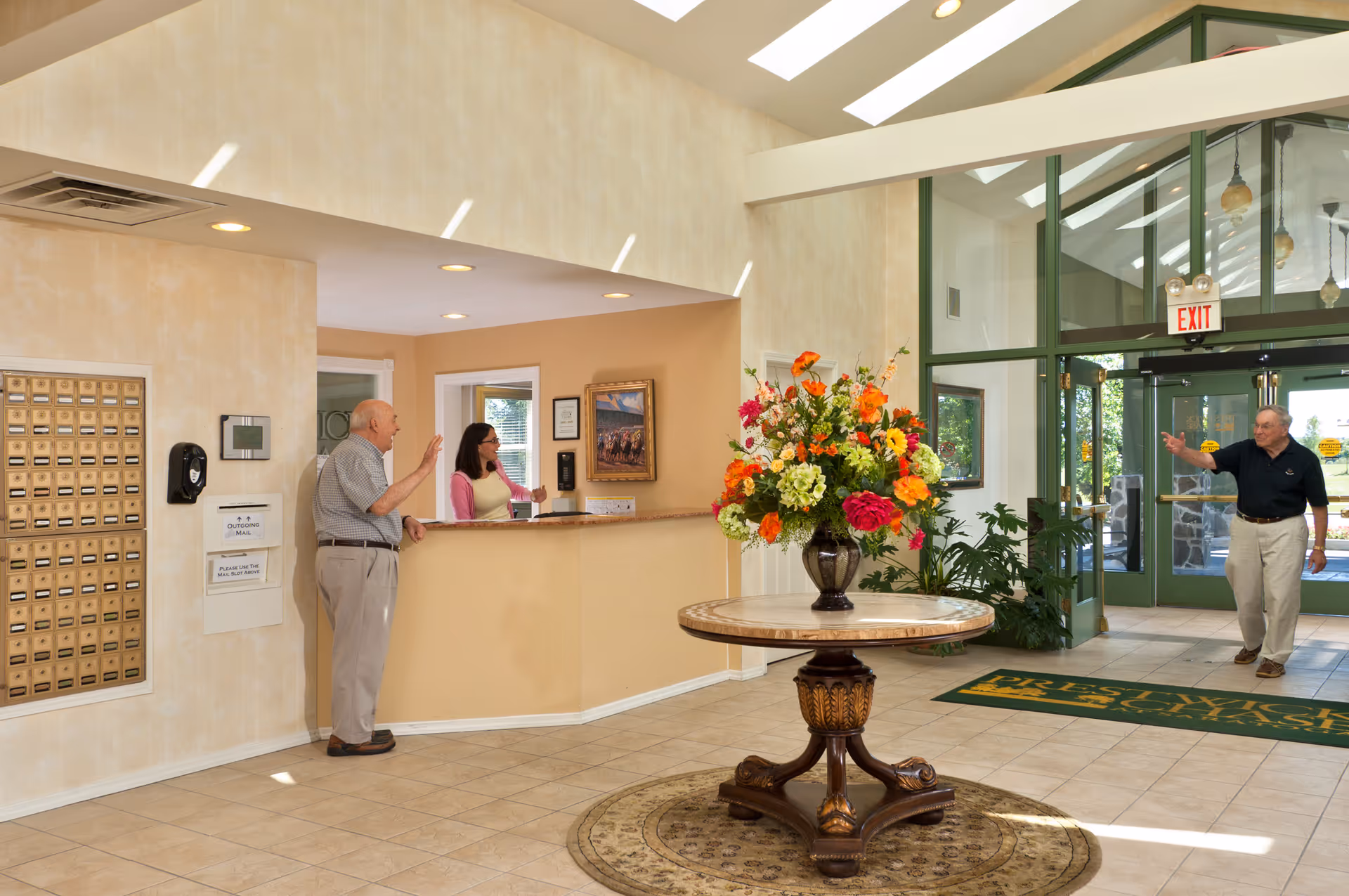 Lobby area of Prestwick Chase at Saratoga featuring a round table with a large floral arrangement in the center. An elderly man is speaking to a receptionist behind the front desk, and another elderly man is entering through glass doors. The space is well-lit with natural light and has beige walls and tiled floors.