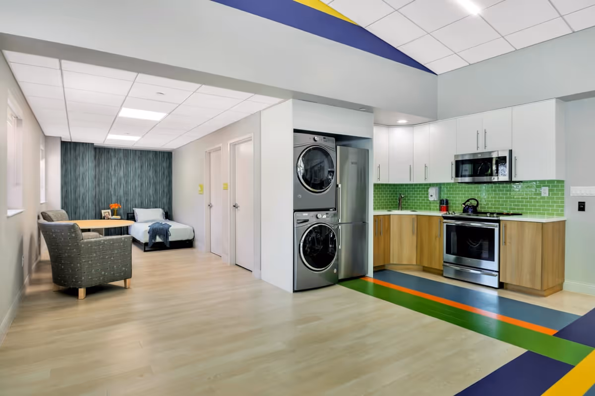 A modern, open interior space featuring a small kitchen area with stainless steel appliances, including a stove, microwave, refrigerator, and stacked washer and dryer. The kitchen has light wood lower cabinets, white upper cabinets, and a green tiled backsplash. Adjacent to the kitchen is a seating area with two armchairs and a small table, and a single bed against a dark textured accent wall. The floor is light wood with colorful stripes near the kitchen area, and the ceiling has recessed lighting and a decorative blue and yellow design.