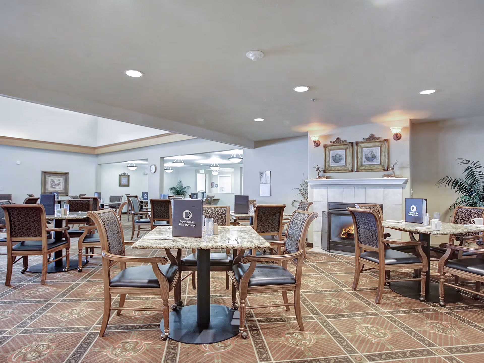 A spacious dining room with multiple tables and chairs arranged neatly. Each table has a menu card and some glassware. The room features a fireplace with two framed pictures above it, and there are plants and wall sconces adding to the decor. The floor has a patterned carpet, and the ceiling has recessed lighting.