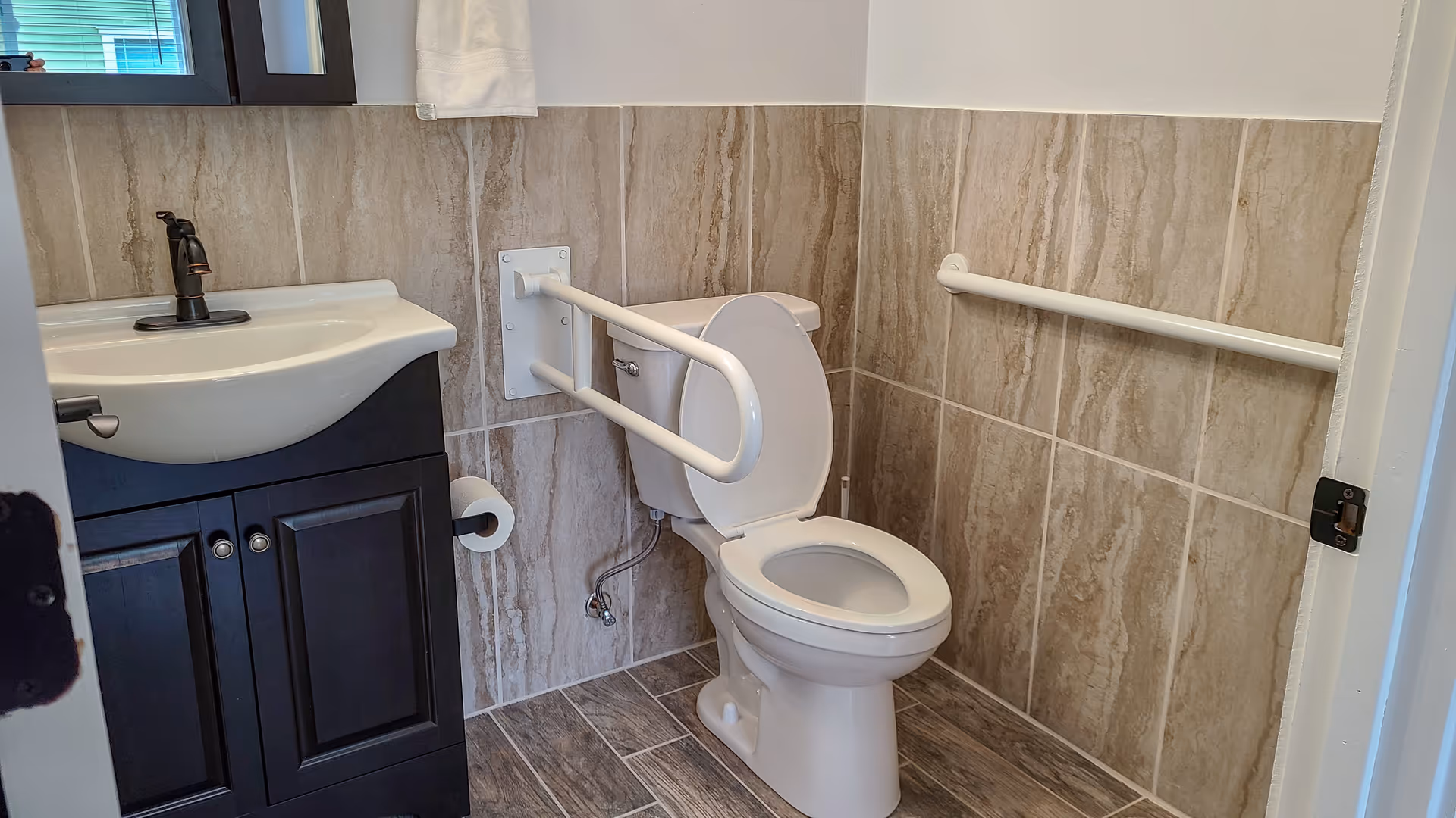 A small bathroom with beige tiled walls and floor, featuring a white toilet with safety grab bars on both sides and a white sink with a dark wooden cabinet underneath. There is a window above the sink with a white towel hanging nearby.