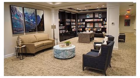 A cozy living room area with beige carpet, a beige sofa, two dark blue armchairs, a round glass coffee table with a plant on it, and a side table with a lamp. The back wall features built-in dark wood bookshelves filled with books and decorative items. Three large landscape paintings hang on the left wall.