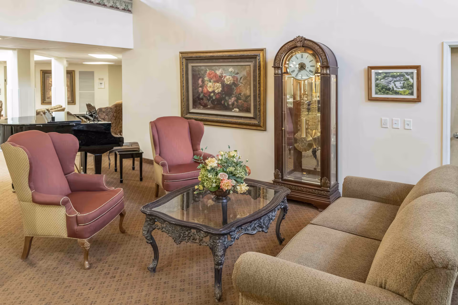 Cozy common living area with upholstered chairs, a sofa, ornate coffee table with flowers, a grandfather clock and a piano in the background.