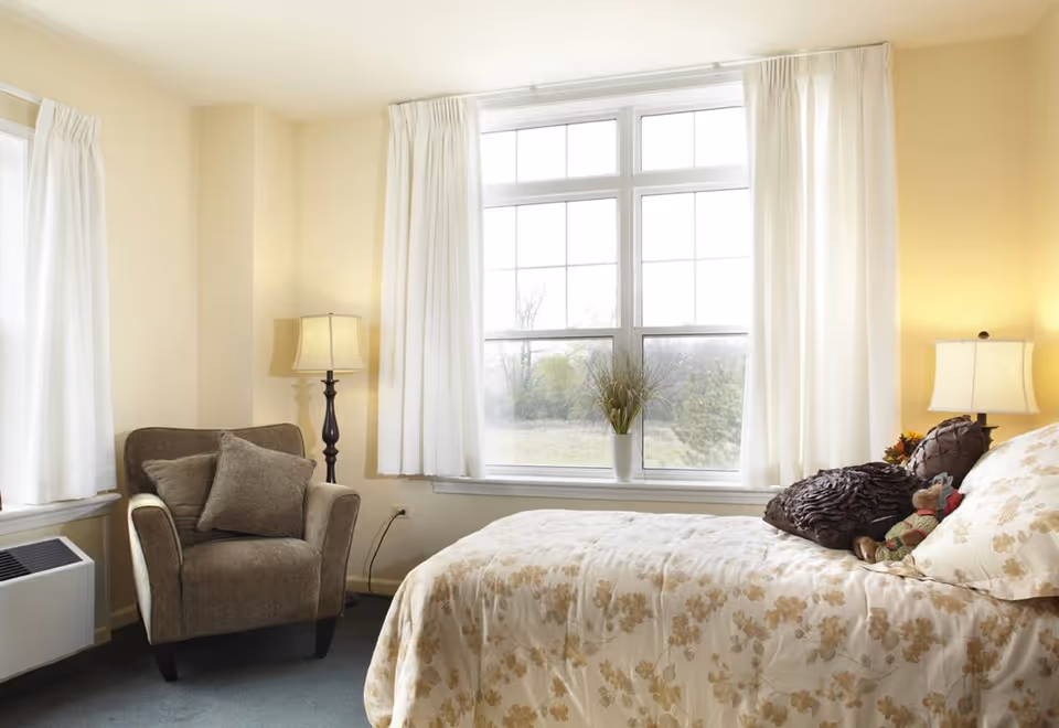 A cozy bedroom with a large window covered by white curtains letting in natural light. The room features a bed with floral-patterned bedding, decorative pillows, and a small stuffed toy. Next to the bed is a nightstand with a lamp and a small flower arrangement. There is also a comfortable armchair with cushions and a floor lamp beside it.