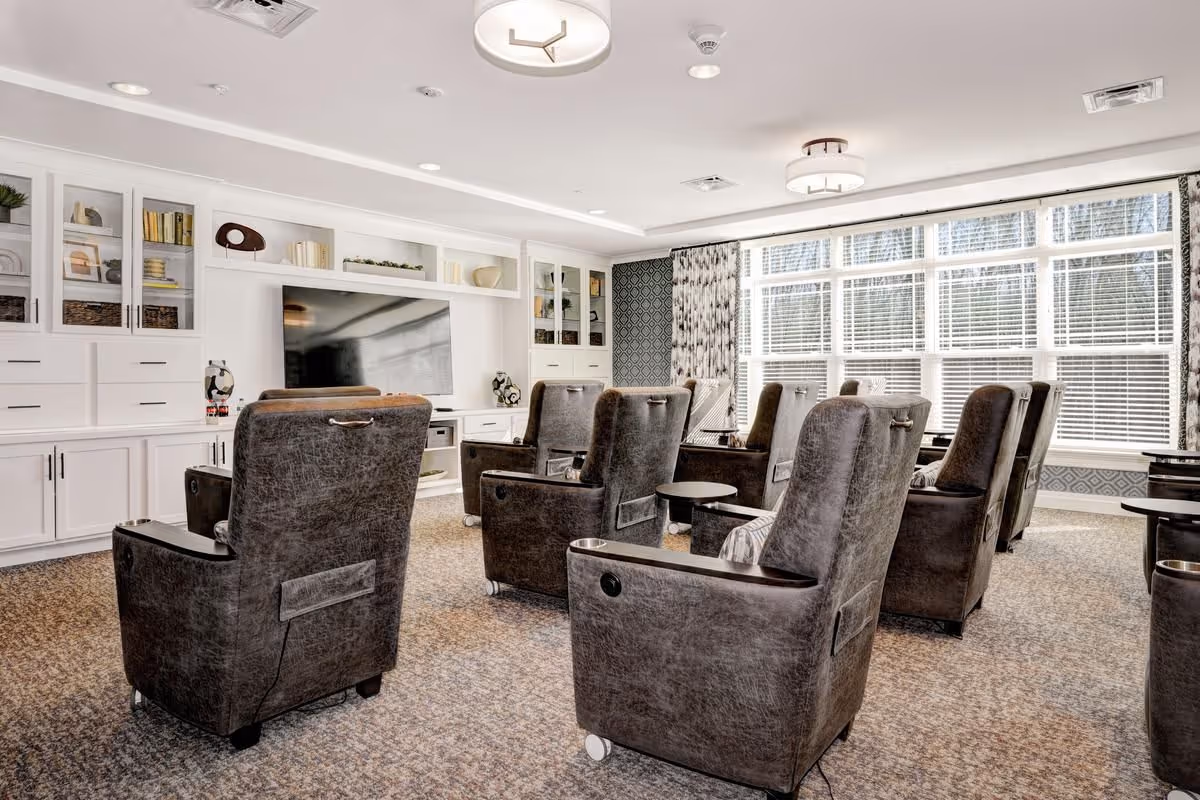 Sunlit communal lounge with rows of leather recliners facing a large wall-mounted TV and built-in white shelving.