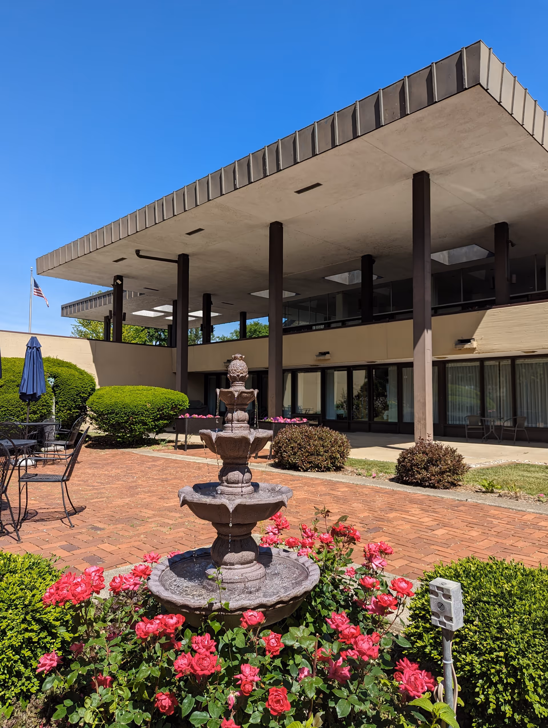 Outdoor patio area at Plum Landing Retirement Community featuring a three-tiered stone fountain surrounded by blooming pink roses and green bushes. The patio has brick flooring with black metal tables and chairs, some with blue umbrellas. The building has a modern design with large overhanging concrete roof supported by dark pillars, and large windows along the wall. An American flag is visible in the background against a clear blue sky.