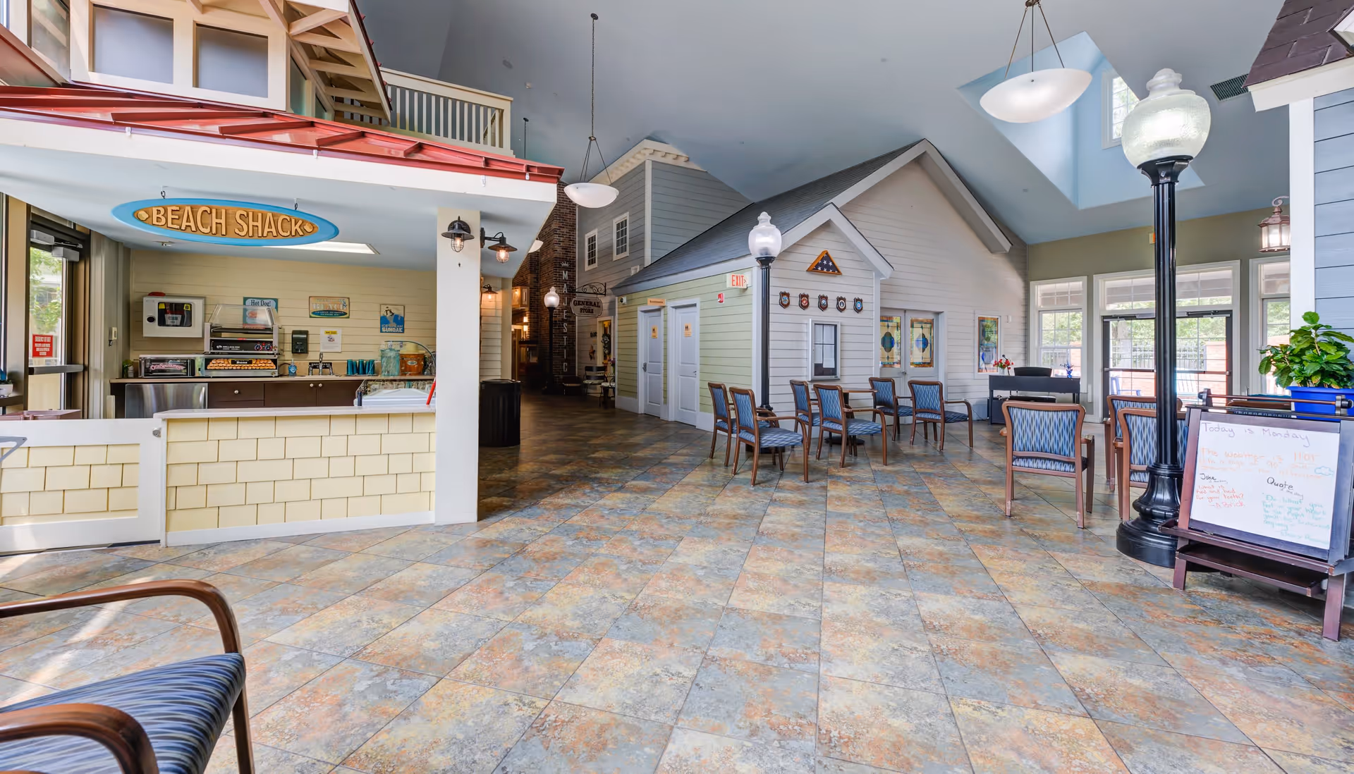 Interior view of a senior living facility common area with a small snack bar labeled 'Beach Shack' on the left, several chairs and tables arranged in the center, and a house-like structure with restroom signs in the background. The space has tiled flooring, high ceilings with skylights, and large windows letting in natural light.