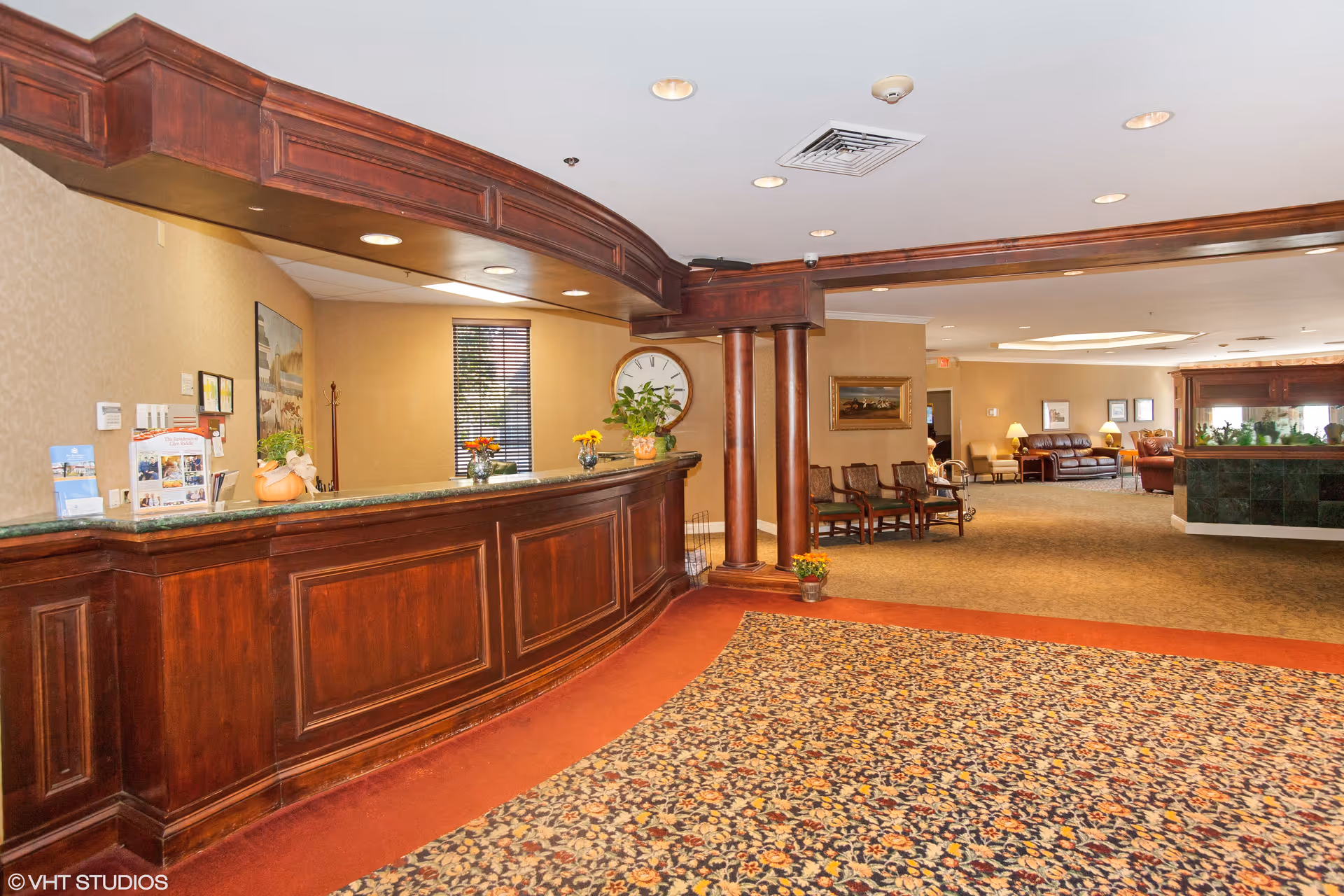 Reception area with a large wooden front desk, floral carpet, and seating area with chairs and sofas in the background. The walls are beige, and there are decorative plants and flowers on the desk. A large clock and framed artwork are visible on the walls.