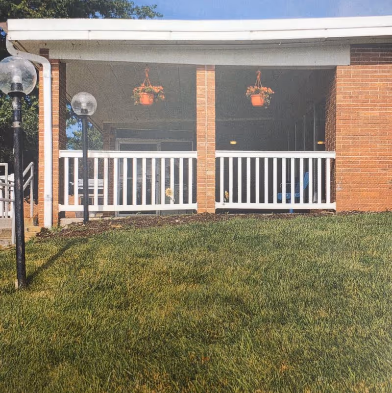 A covered porch area with white railing and brick pillars at Prestige Care Center of Plattsmouth. Two hanging flower pots with orange flowers are suspended from the porch ceiling. There are two lamp posts with round glass tops on the left side near the steps leading up to the porch. The foreground shows a well-maintained green lawn.