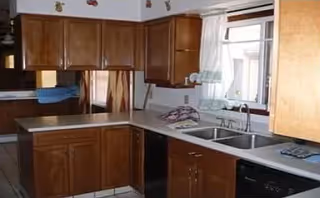 A kitchen with wooden cabinets, a double sink under a window with white curtains, a dishwasher, and a countertop with various items including a dish towel and a lamp.