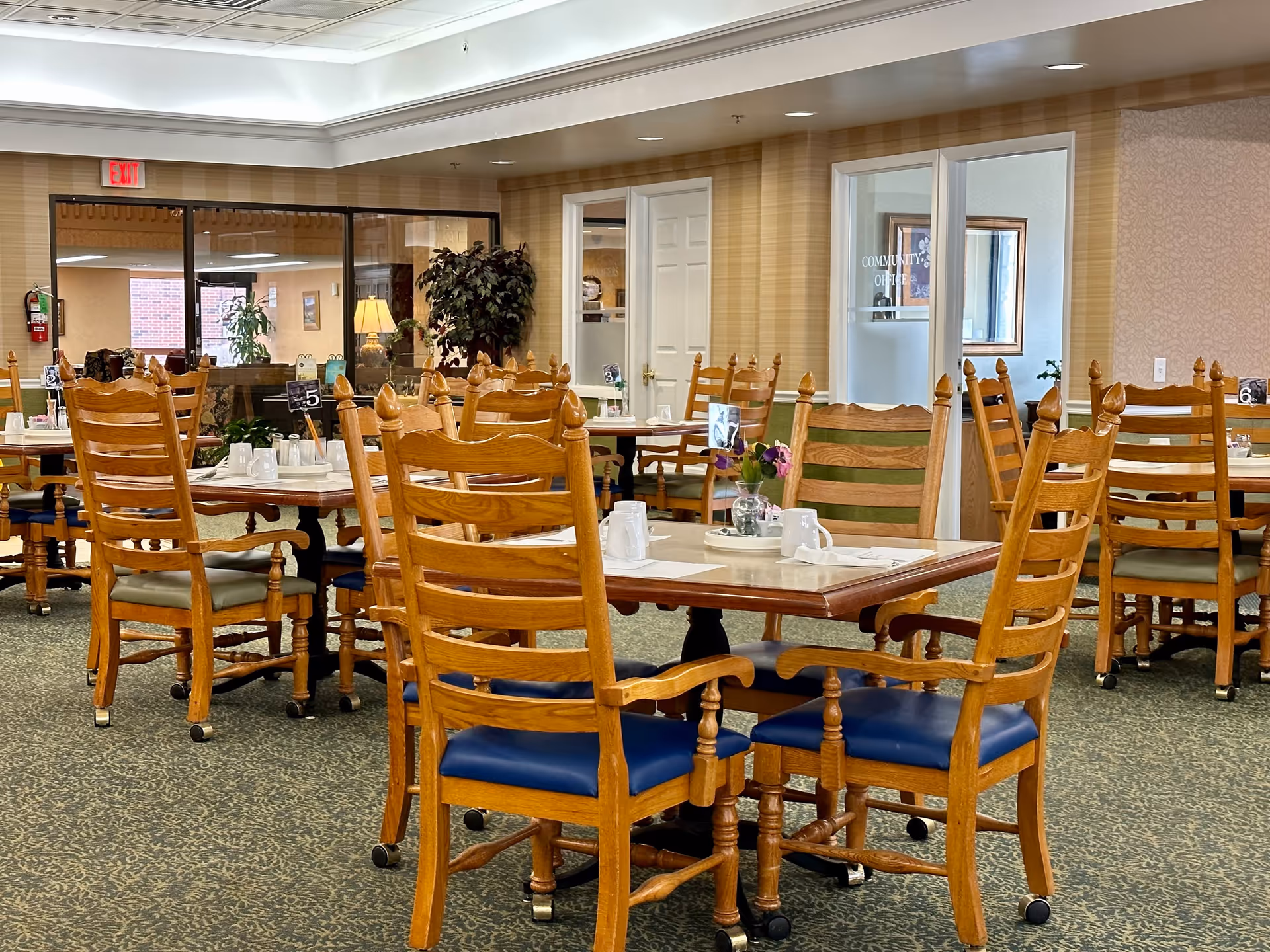 A dining room with multiple wooden tables and chairs with blue cushions. Each table is set with white mugs, napkins, and small flower arrangements. The room has patterned carpet, beige striped wallpaper, and a glass door labeled 'Community Office'. There is an exit sign above a door in the background.