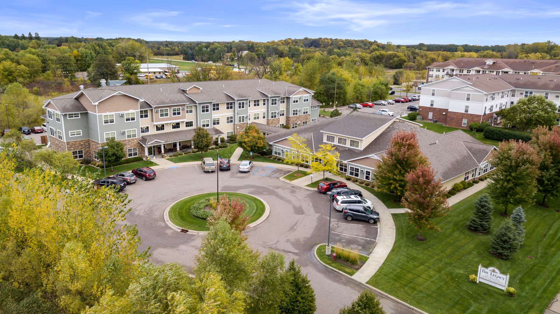 Aerial view of Legacy of Delano Senior Living facility showing multiple connected buildings with a circular driveway and parking spaces. The surrounding area has green lawns, trees with autumn foliage, and a sign for the facility near the entrance.