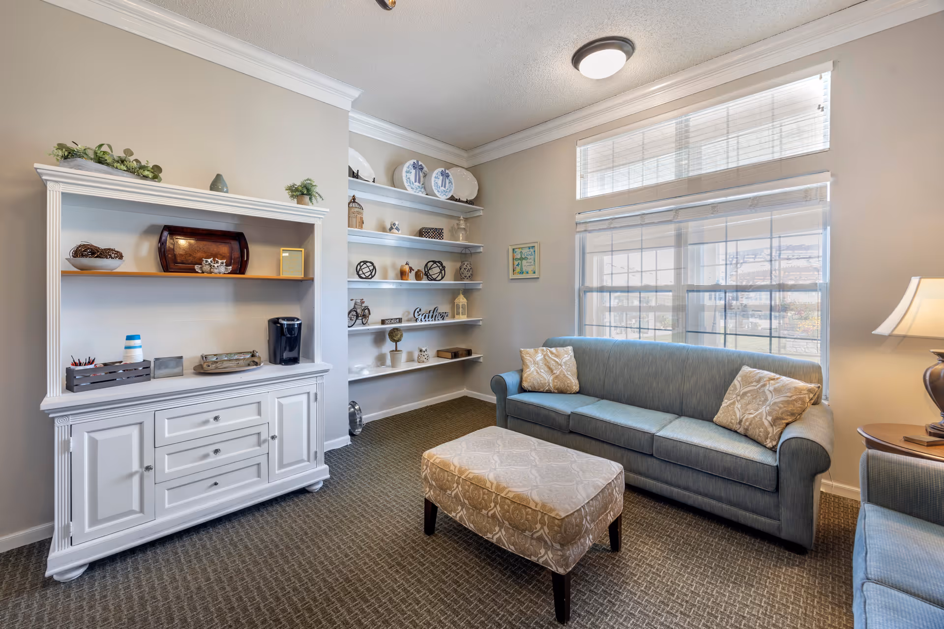 A cozy living room with a blue sofa, matching armchair, and a patterned ottoman in the center. There is a white cabinet with shelves holding decorative items and a coffee maker on the left side. Built-in white shelves with various decorative pieces are on the back wall. Large windows with blinds allow natural light to fill the room. A table lamp is visible on the right side.