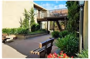 Outdoor courtyard area with benches, raised plant beds, and various green plants and flowers under a clear blue sky. There is a staircase leading to an upper level balcony attached to a beige building.