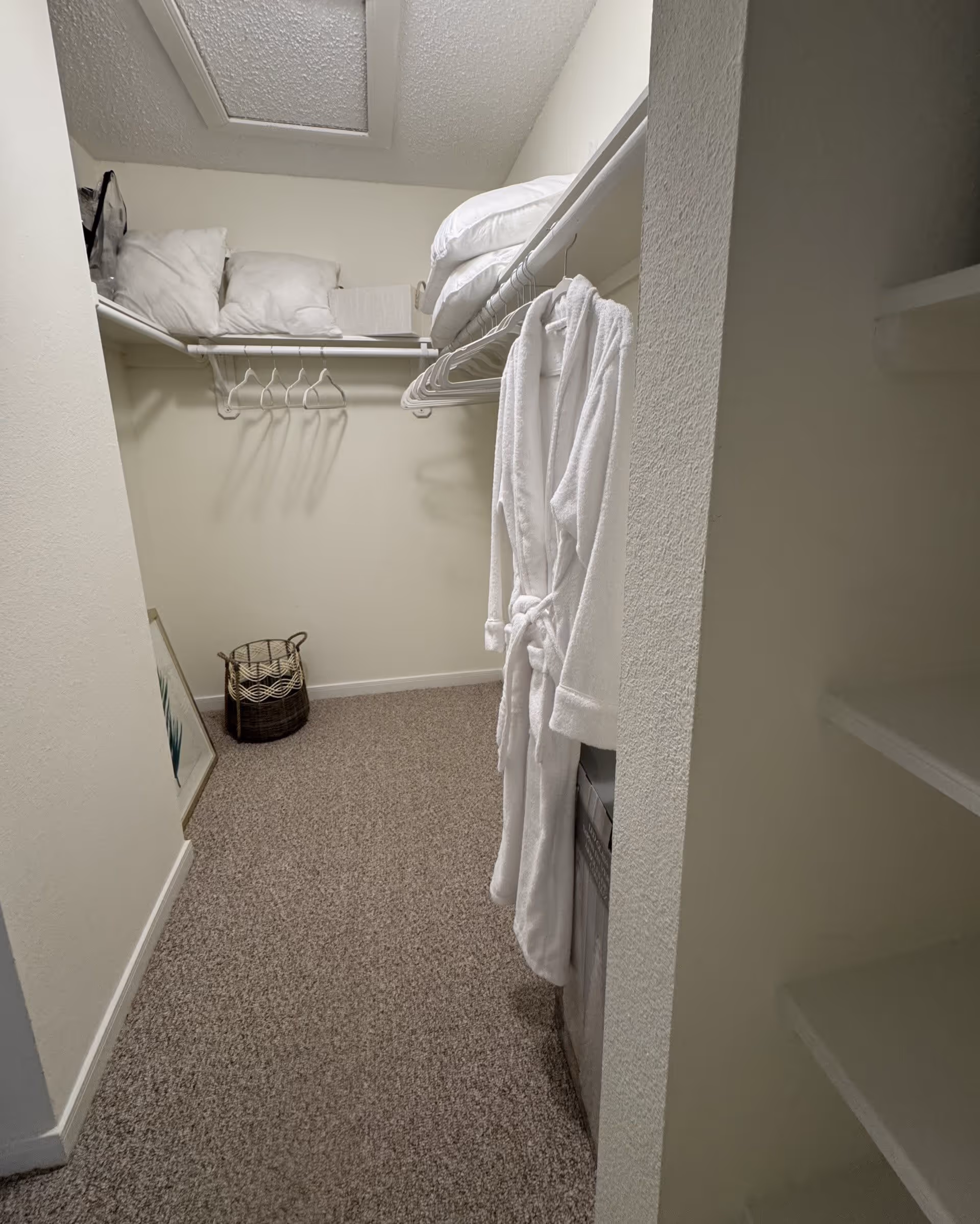 Walk-in closet with beige carpet and white walls, featuring hanging white bathrobes, empty hangers, pillows and blankets on the upper shelf, a woven basket on the floor, and some shelving on the right side.