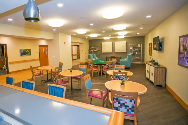 A bright and spacious common area in a nursing home with several round wooden tables surrounded by colorful chairs. In the background, there are two teal armchairs and a fireplace with decorative shelves on either side. The walls are decorated with framed artwork, and a flat-screen TV is mounted on the right wall. The floor is dark wood, and the ceiling has multiple round light fixtures.