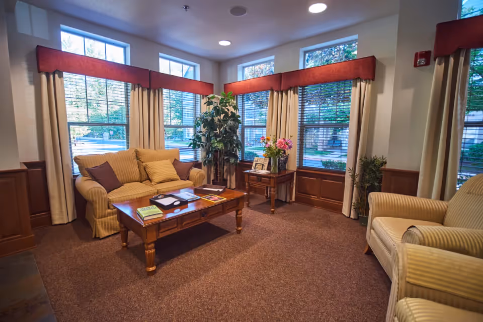 A cozy living room area with a beige striped sofa and armchair, a wooden coffee table with books and a tray, a side table with a vase of flowers, and large windows with beige curtains and red valances letting in natural light.