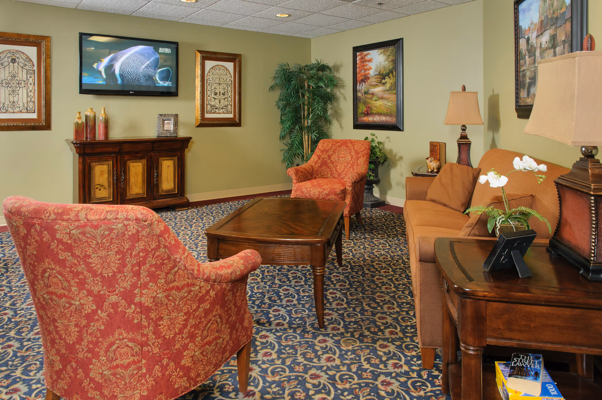 Cozy sitting area with patterned armchairs, a sofa, wooden coffee and side tables, a wall-mounted TV, framed artwork and a patterned carpet.
