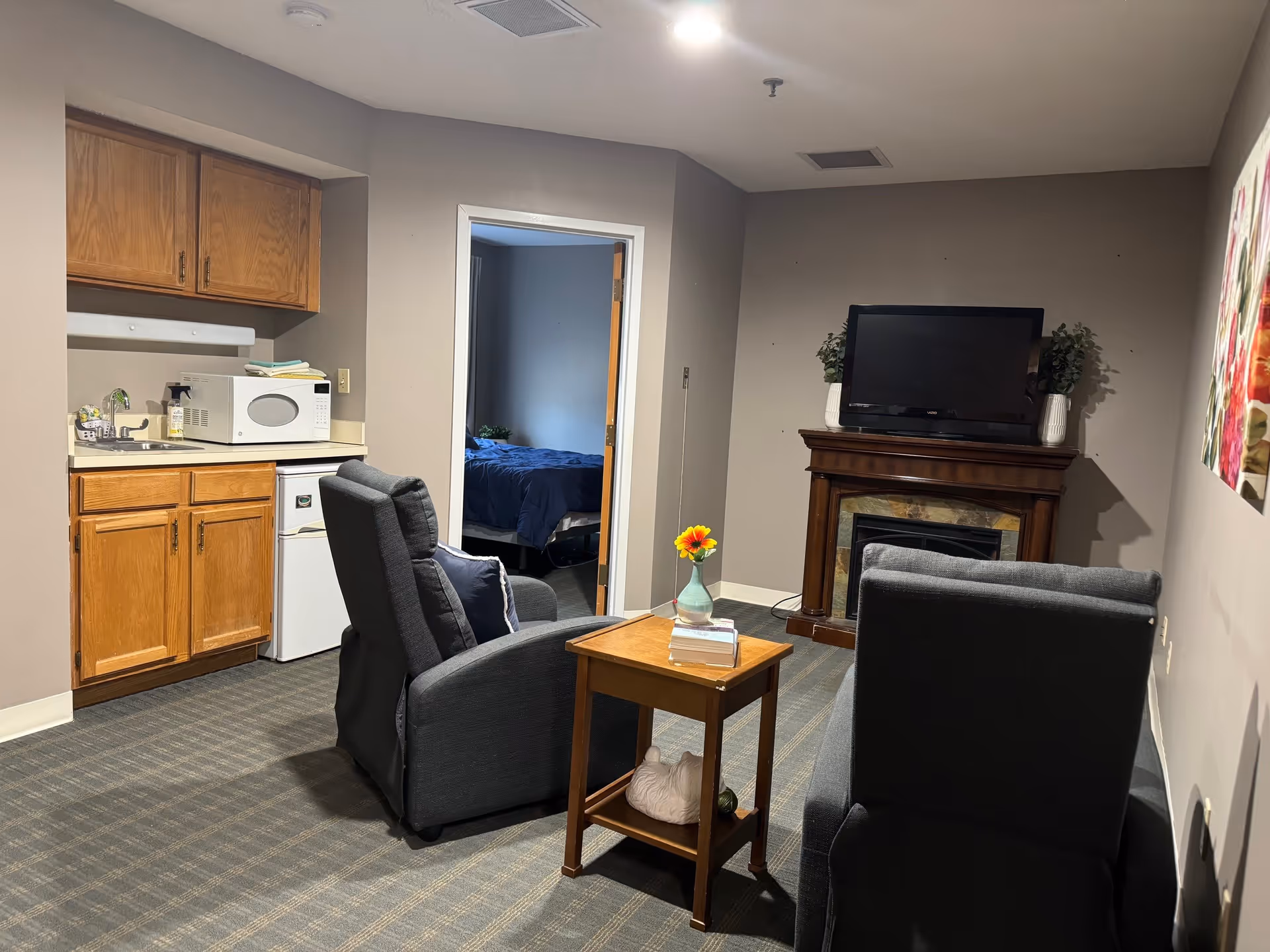 Small senior living suite interior with a kitchenette, two armchairs facing a TV on a fireplace mantel, and a doorway leading to a bedroom.