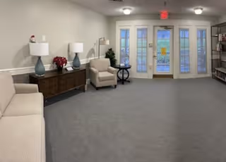 A spacious senior living facility common area with beige walls and carpeted floor. The room features a beige sofa on the left, a wooden sideboard with two blue lamps and a red flower arrangement, a beige armchair next to a small round table, and a bookshelf on the right. Double glass doors with white frames lead outside, and ceiling lights illuminate the room.