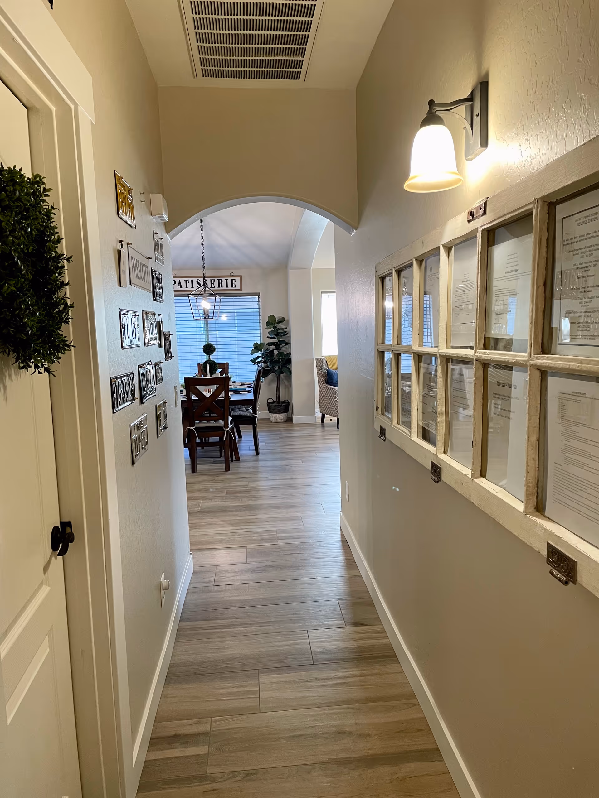 A hallway in a senior living facility with light wood flooring and beige walls. On the left wall, there are decorative plaques and a wreath on the door. On the right wall, there is a framed display with multiple documents behind glass. A wall-mounted light fixture illuminates the hallway. At the end of the hallway, there is an arched opening leading to a dining area with a wooden dining table, chairs, a hanging light fixture, and a large window with blinds. A potted plant is visible near the window.